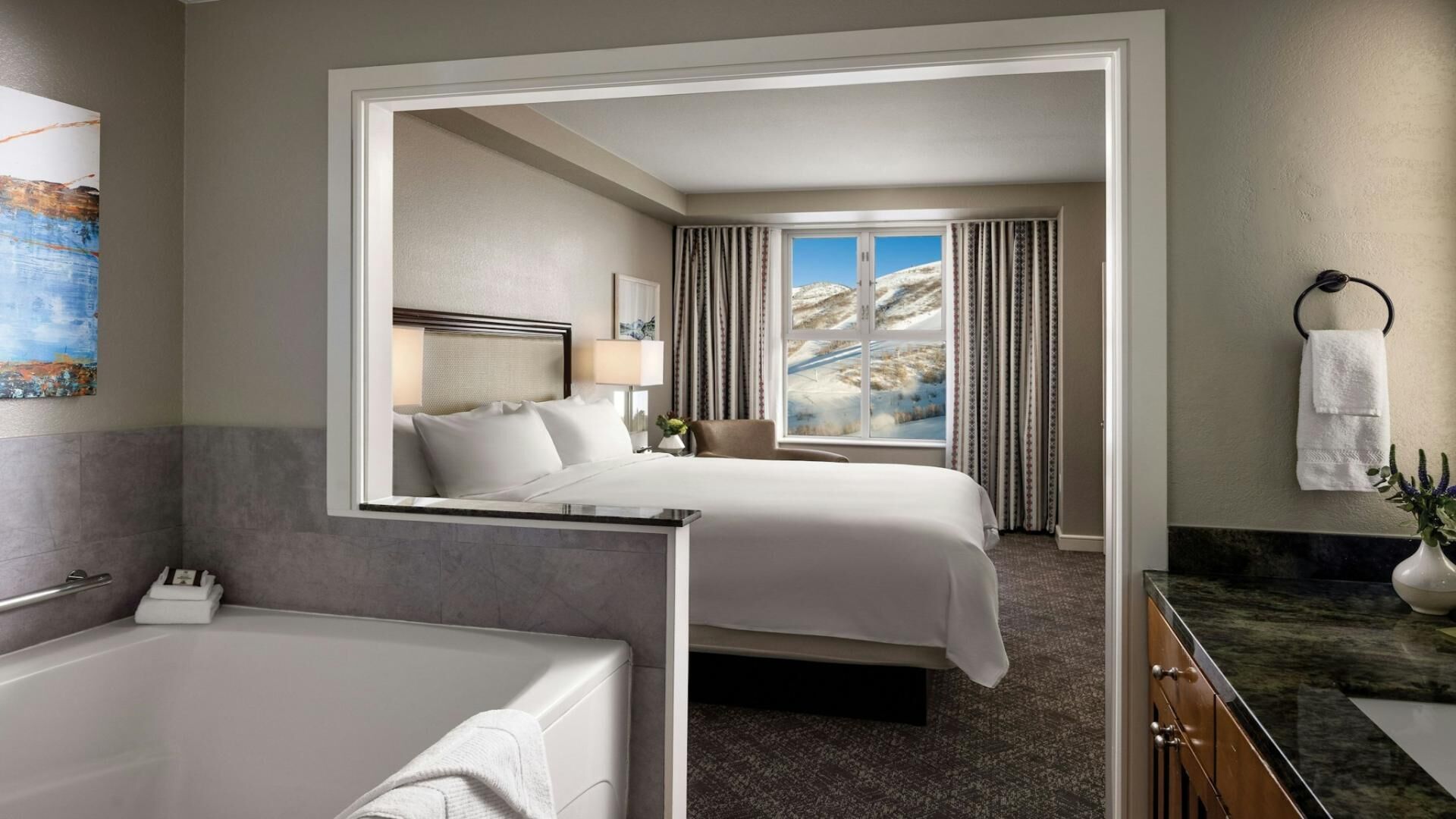 Une chambre d’hôtel avec un lit, une salle de bain dotée d’une baignoire, un miroir et une vue sur un paysage enneigé par la fenêtre.