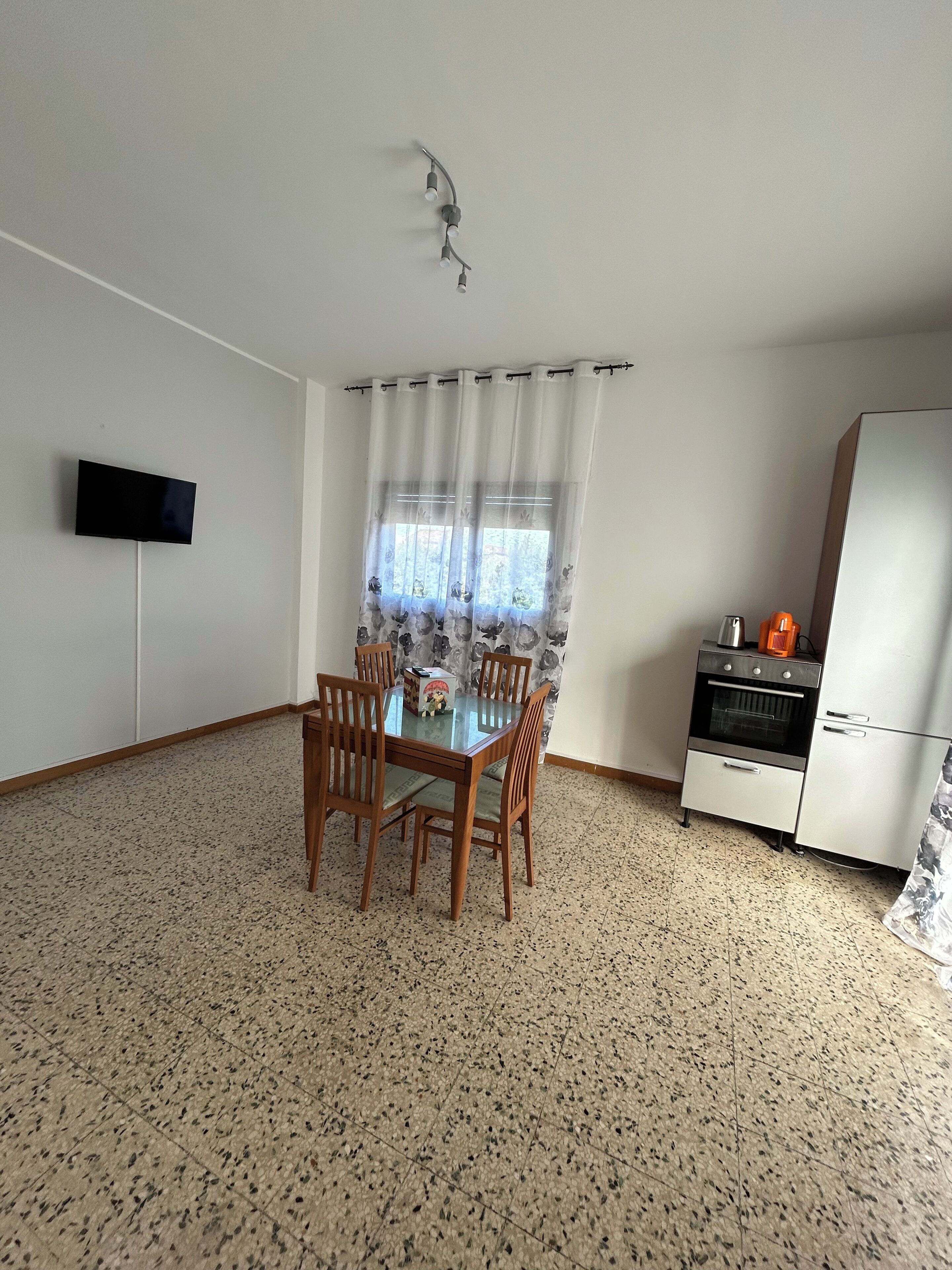 A dining area with a table and chairs, a television mounted on the wall, a window with curtains, and a kitchenette with appliances.