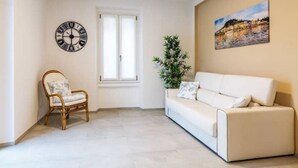A living room with a white sofa, a wicker chair, a clock, a window, and a painting on the wall. - Celia's house near Bellagio (Lezzeno)