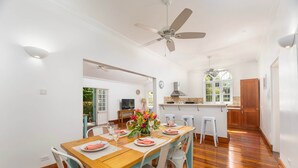 A dining area with a wooden table, white chairs, a ceiling fan, and a kitchen in the background. - Hummingbird Villa (Porters)