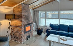 Un salon chaleureux avec une cheminée en briques, un canapé bleu, une table basse et une grande fenêtre donnant sur l’extérieur.
