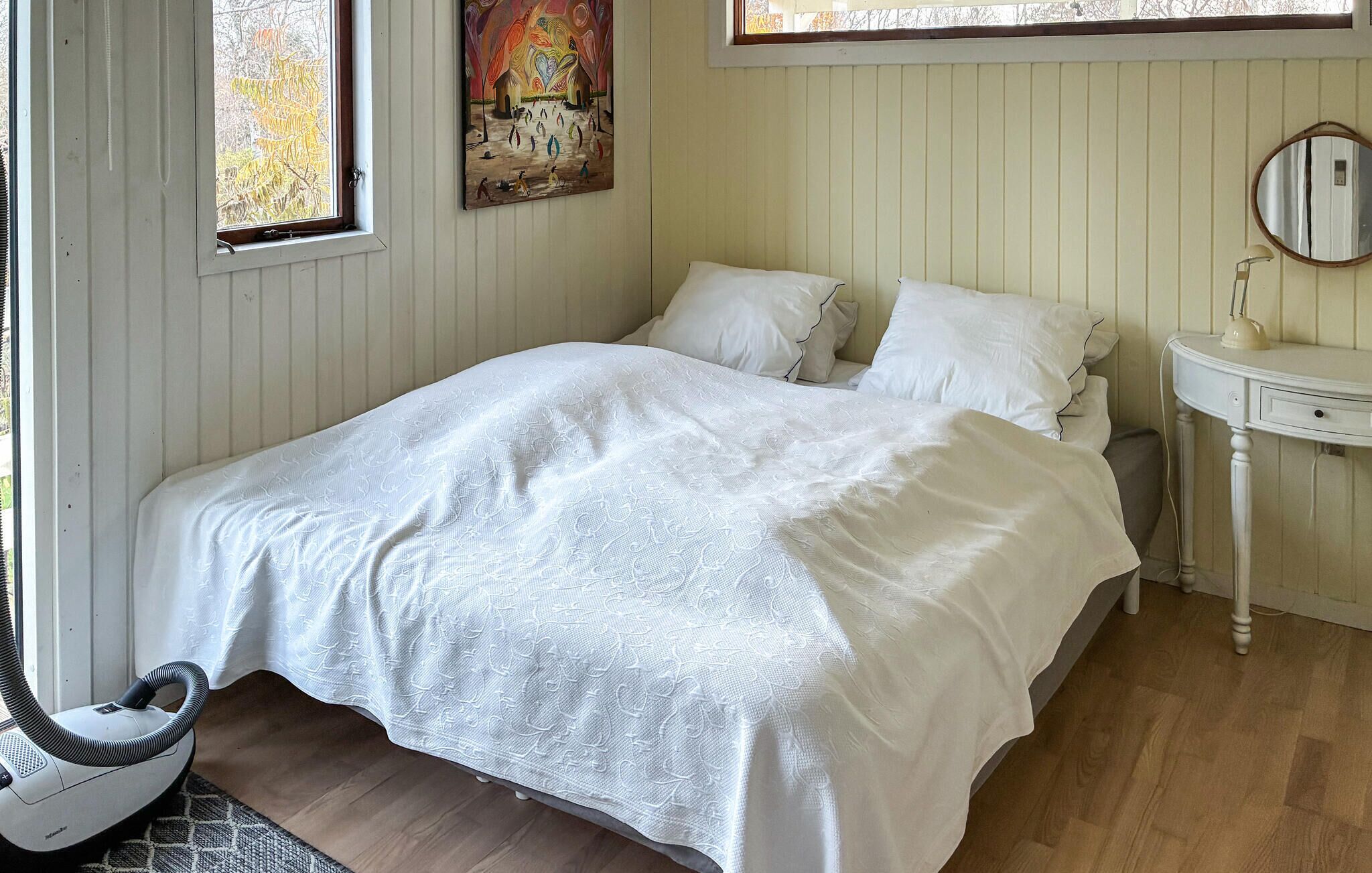 Un dormitorio con cama, mesita de noche, espejo y aspiradora.