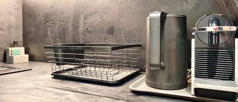 Cozinha com cafeteira de aço inoxidável, cesto de arame e dispensador de sabonete no balcão.