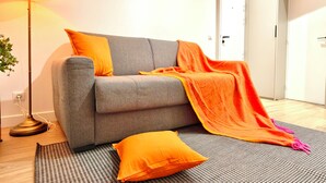 Un divano grigio con cuscini arancioni e una coperta, una lampada da terra e un tappeto decorato in una stanza con una porta e una presa elettrica.
