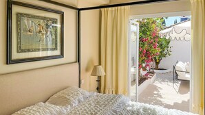 Ein Schlafzimmer mit einem Bett, einem gerahmten Bild an der Wand, einer Lampe und Blick auf einen Garten durch eine Glastür.