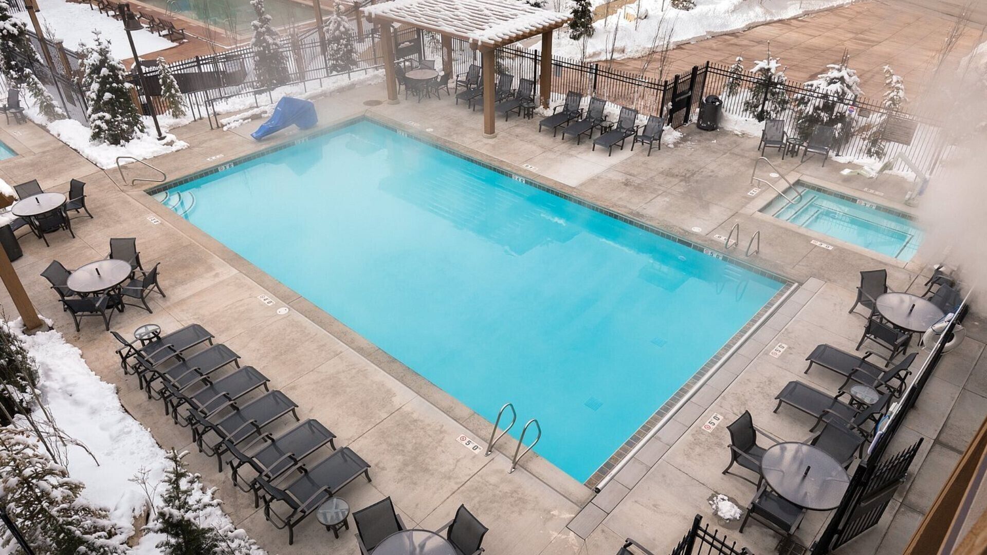 Una piscina rectangular con un tobogán azul, rodeada de sillas de descanso y mesas, con nieve en el suelo.