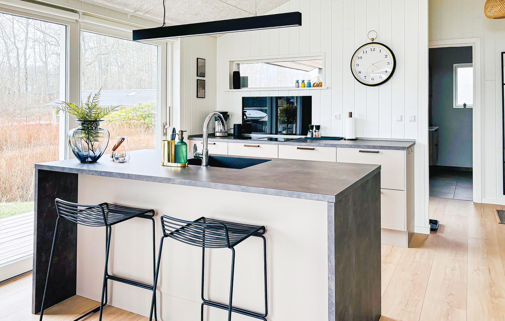 A modern kitchen with a central island, bar stools, a clock, and a view of the outdoors.