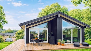 A modern house with a wooden deck, a dining area with chairs and a table, and a large glass door leading to a covered outdoor space.