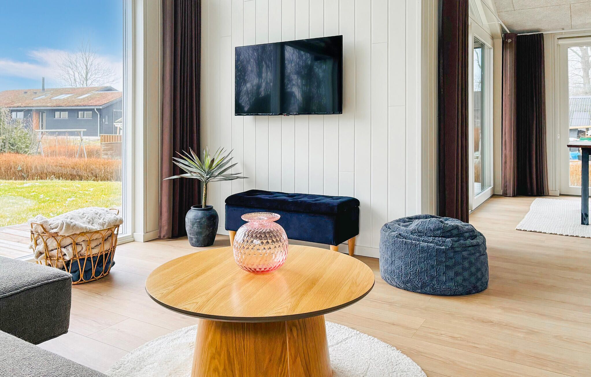 A modern living room with a round wooden table, a dark blue bench, and a blue bean bag. A flat-screen TV is mounted on the wall, and a potted plant is on the floor.