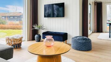A modern living room with a round wooden table, a dark blue bench, and a blue bean bag. A flat-screen TV is mounted on the wall, and a potted plant is on the floor.