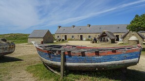 Ein traditionelles Steinhaus mit einem strohgedeckten Dach, ein Boot im Vordergrund und ein klarer, blauer Himmel.