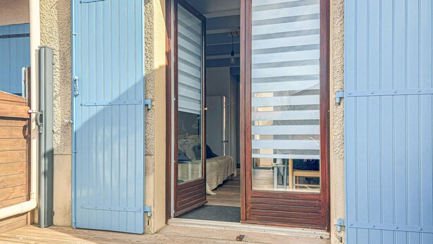 Een houten terras leidt naar een kamer met een glazen schuifdeur en een bed met een beddengoed met een patroon.