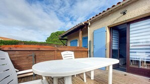 Een terras met een witte tafel en stoelen, een houten terras en een gebouw met blauwe luiken en een schuifdeur van glas.