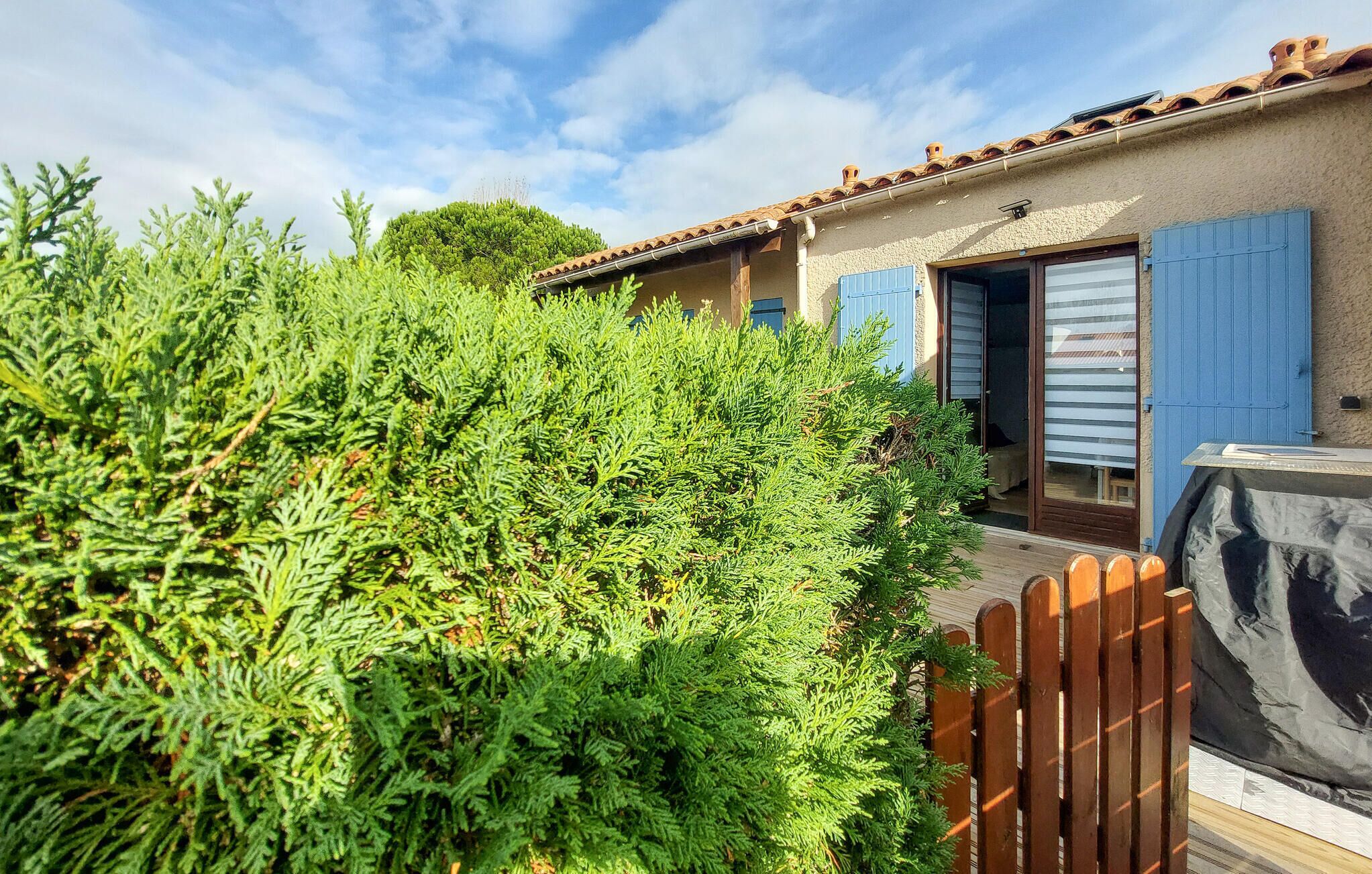 A rustic house with a tiled roof, a wooden fence, and a garden with dense green foliage.