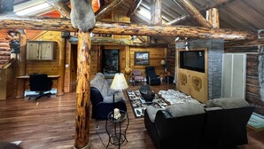 A rustic cabin interior with wooden walls, a stone fireplace, a dining area, and a living room with a sofa and chairs. - Cozy Log Retreat Near Turnagain Arm (Anchorage)