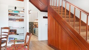 Una escalera con barandillas de madera que sube a un piso superior, una estantería con varios libros y un sillón con un cojín de colores.