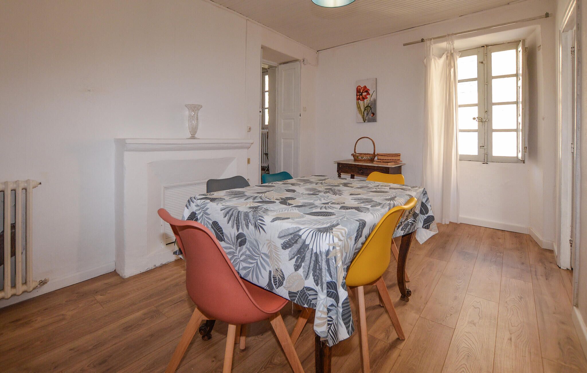 A dining area with a table set for four, featuring a patterned tablecloth. There are two chairs, one red and one yellow, and a small table with a basket and a vase. A radiator is visible on the left, and a door with a window is on the right.
