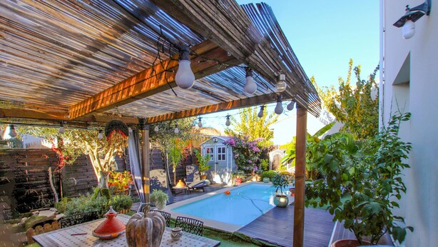 Un espacio exterior techado con piscina, pérgola de madera y diversas plantas en macetas.