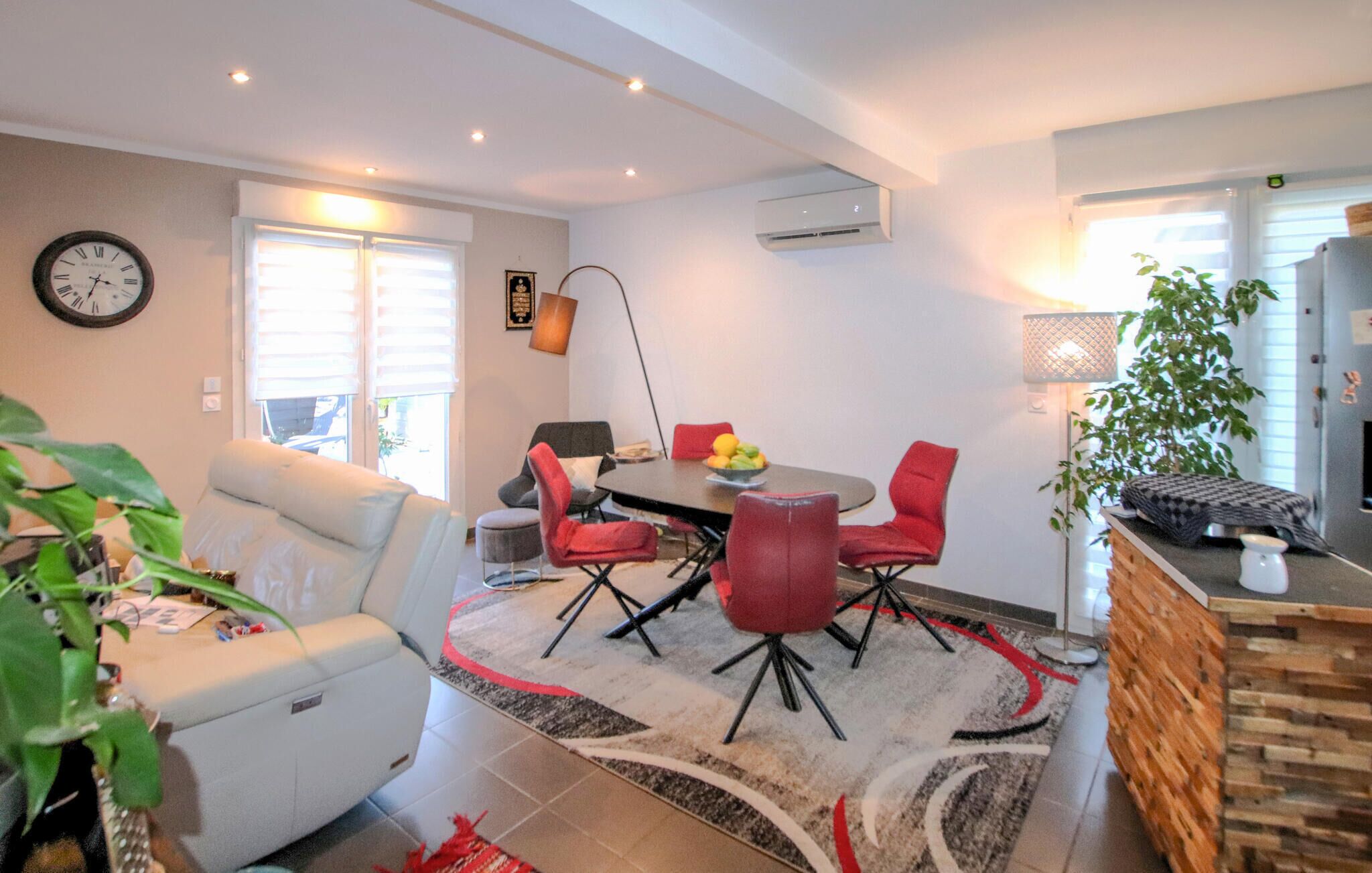 A modern living room with a sofa, a dining table with red chairs, a clock on the wall, and a plant.
