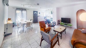 Un vaste espace de vie comprenant une table à manger, plusieurs chaises, un canapé, un téléviseur et, au fond, une cuisine.