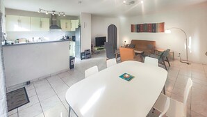 Une cuisine moderne avec des meubles blancs, un coin repas avec une table et des chaises blanches, et un espace de vie avec un canapé et un téléviseur.