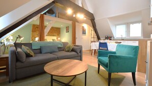 Ein Wohnzimmer im Loft-Stil mit einer Couch, einem türkisfarbenen Sessel und einem runden Tisch. Eine Treppe führt zu einer oberen Ebene, auf der eine Küche zu sehen ist.