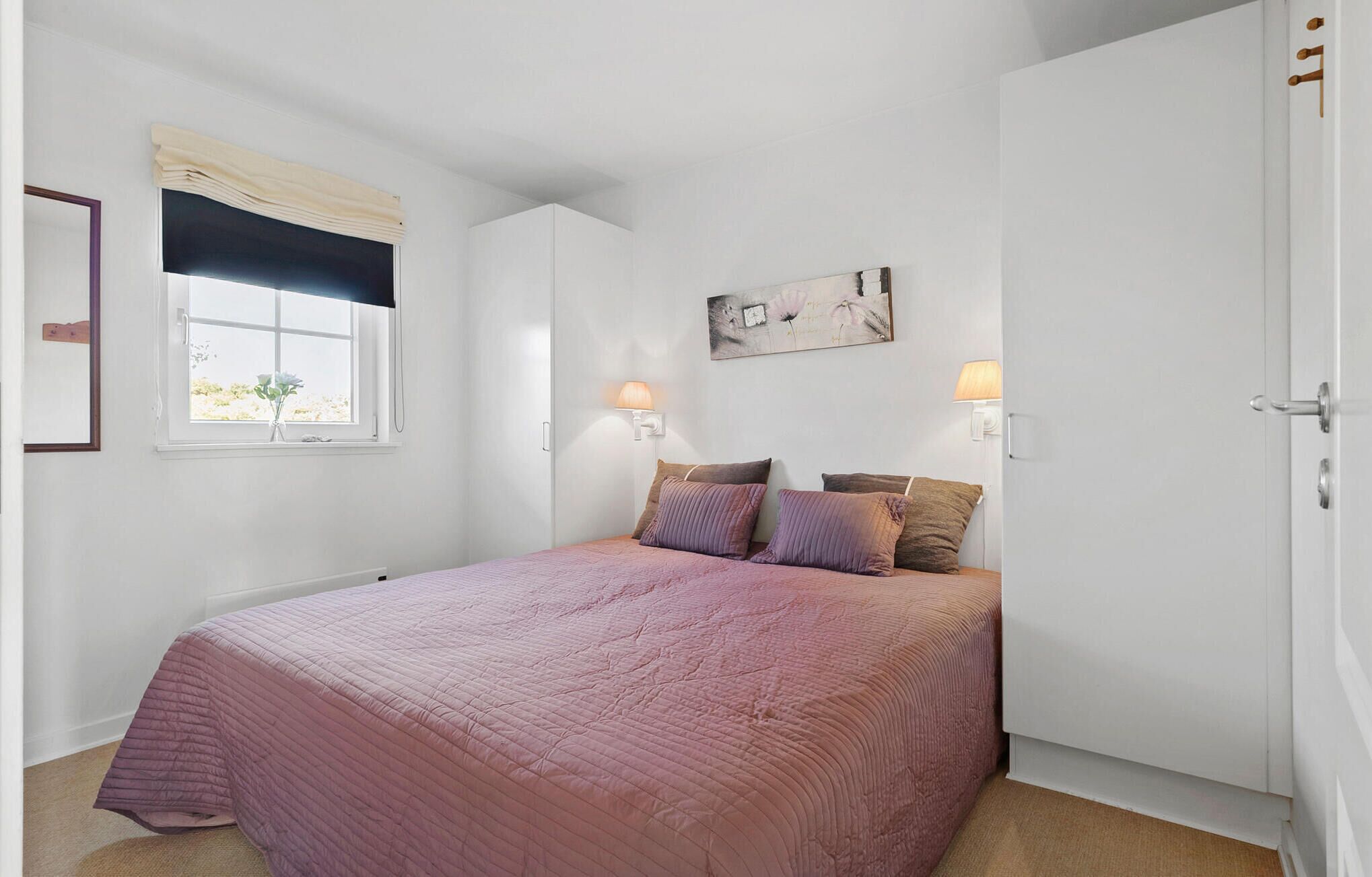 Ein Schlafzimmer mit einem Bett, zwei Nachttischlampen, einem Fenster mit Vorhang, einem Bild an der Wand und einer Tür.