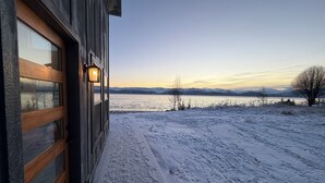 Una cabana de fusta amb una porta de vidre i un llumí encès, amb vista a un paisatge nevat que inclou un llac i muntanyes a la llunyança.