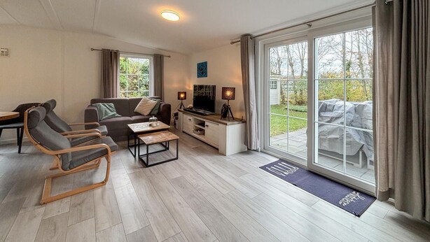 Ein modernes Wohnzimmer mit einer Couch, Sesseln, einem Couchtisch und einem Fernseher. Eine Glastür öffnet sich zu einer Terrasse mit Gartenmöbeln.