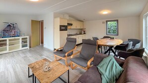 Ein modernes Wohnzimmer mit einer Couch, Sesseln und einem Couchtisch. Im Hintergrund ist eine Küche mit Schränken und einem Fenster zu sehen.