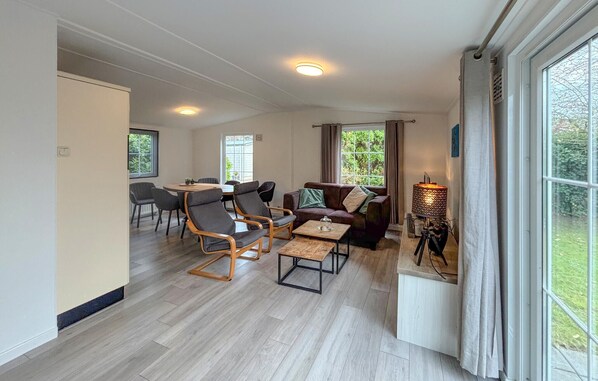A spacious living area with a dining table, comfortable seating, and large windows offering a view of greenery. - Awesome (sta)caravan in Goedereede with WiFi (Goedereede)
