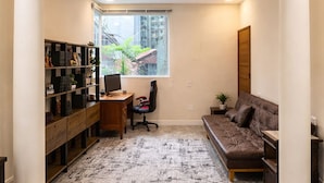 A room with a desk, chair, bookshelf, sofa, and large windows.