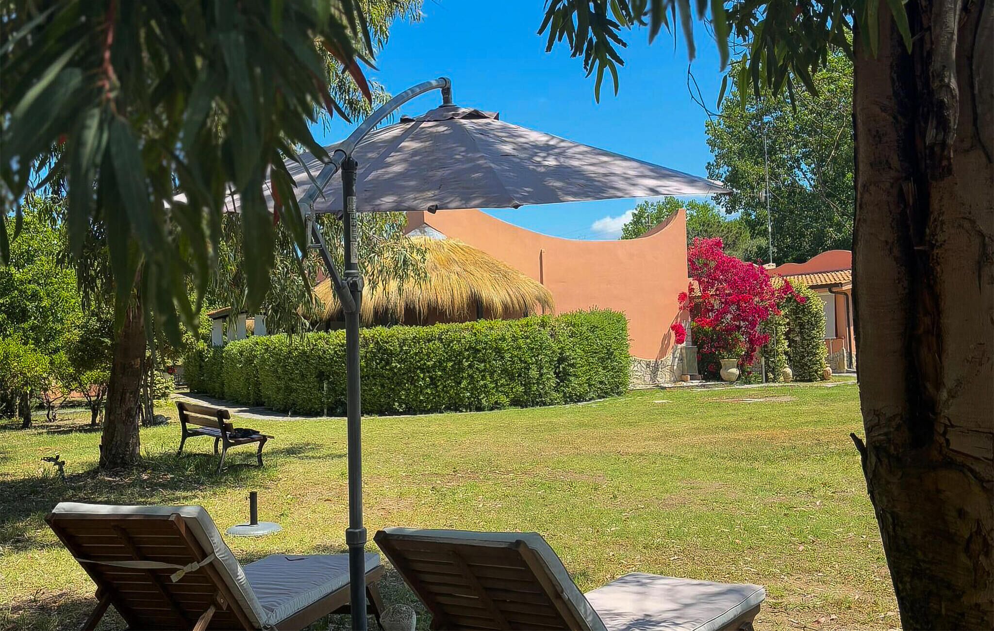A patio area with lounge chairs and a large umbrella, surrounded by well-maintained hedges and a thatched-roof structure.