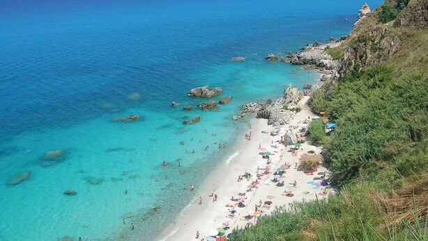 Een strand met helder turquoise water, rotsformaties en een zandstrand met parasols en mensen.