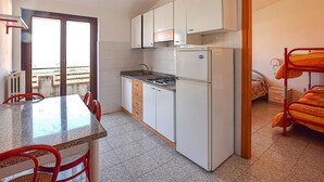 Een kleine keuken met witte kasten, een gasfornuis en een koelkast. Er is een zithoek met een tafel en stoelen, en een stapelbed met oranje beddengoed.