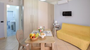 Een moderne woonkamer met een houten eettafel, gedekt met fruit en bloemen, een gele bank en een televisie aan de muur.