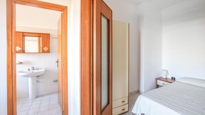 Een slaapkamer met een bed, nachtkastje met lamp, kast en een badkamer met wastafel en spiegel.