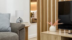 A modern living room with a grey sofa, a small side table with a lamp, a wooden TV stand with a flat-screen TV, and a mirror reflecting the room. - Chueca Center II - Studio for 4 in downtown Madrid (Madrid)