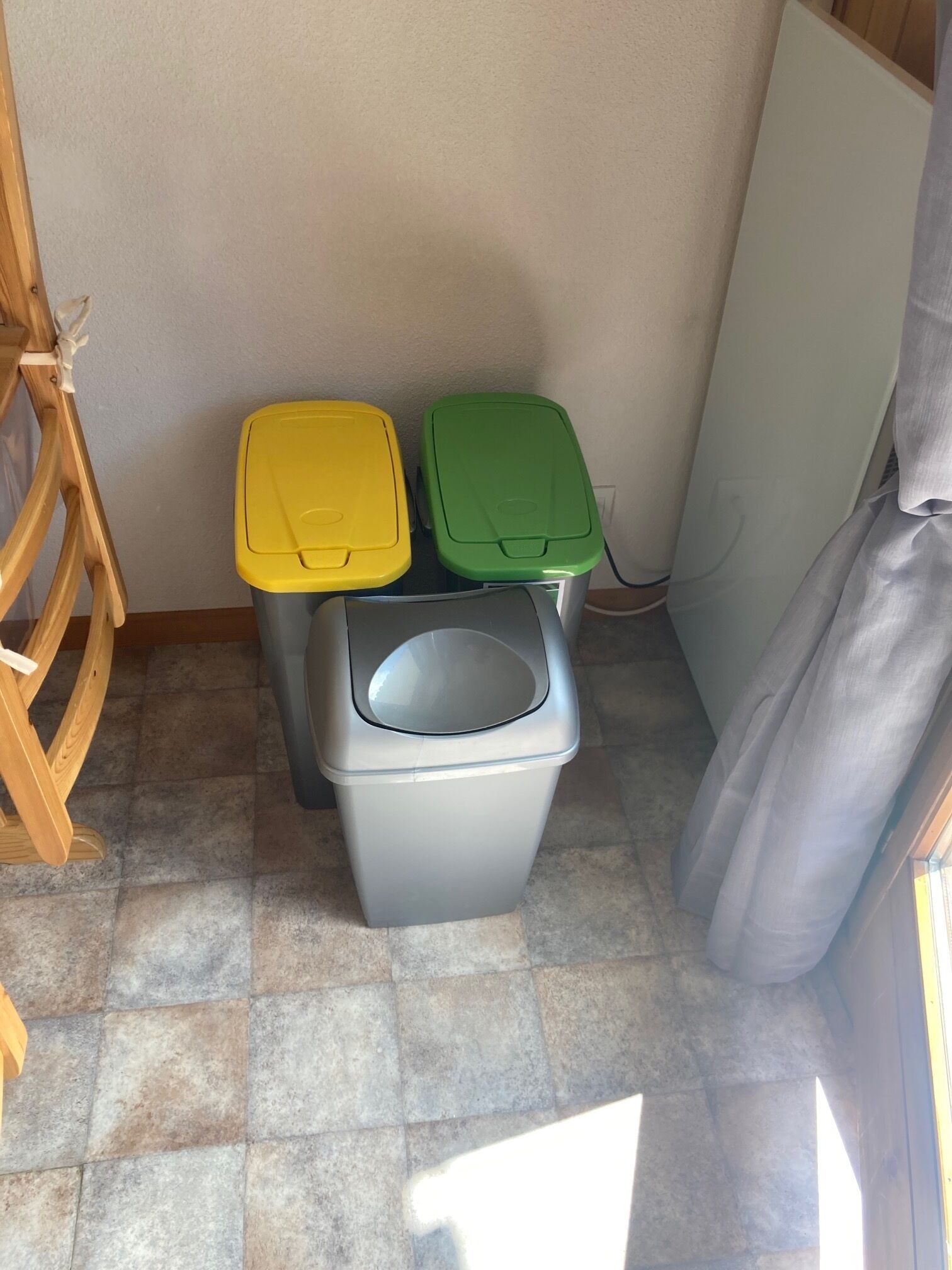 A corner with three trash bins in different colors and a wooden chair.