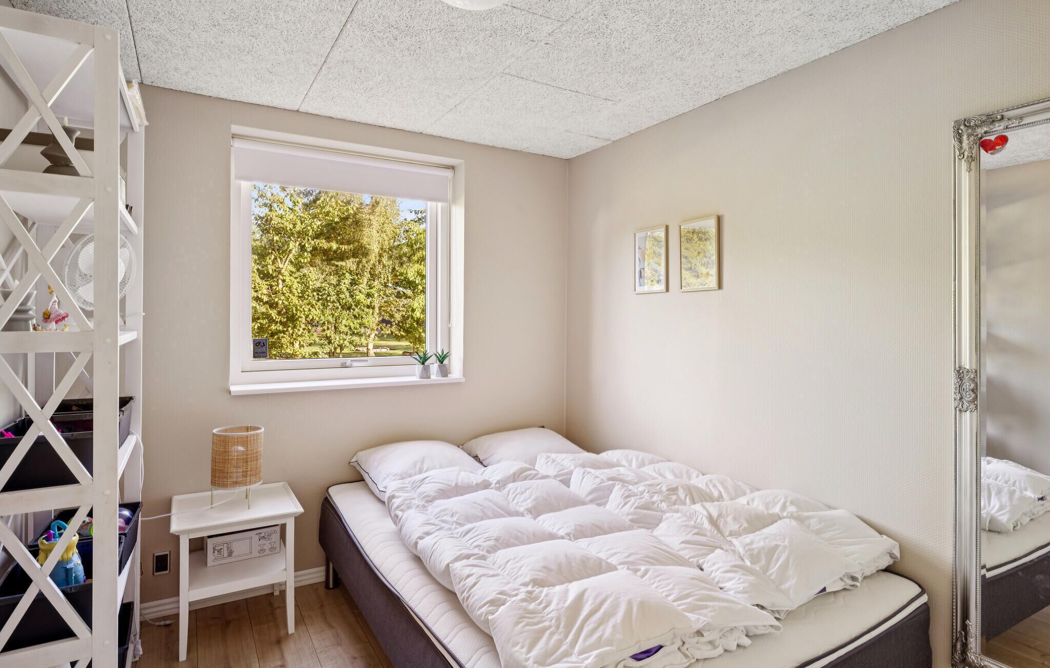 Une chambre à coucher avec un lit, une fenêtre donnant sur des arbres, une table de chevet et un miroir.