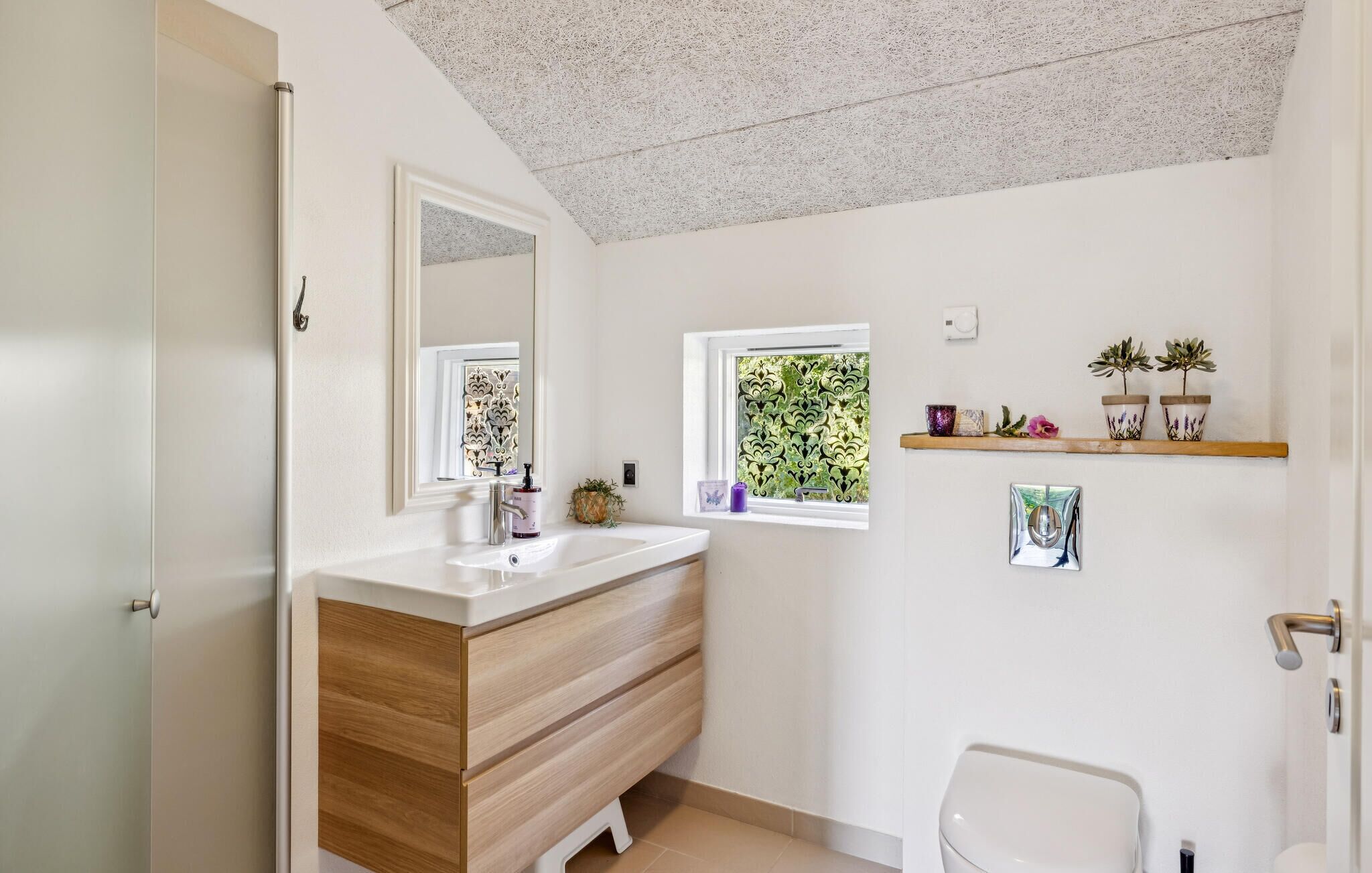 A bathroom with a white sink, wooden vanity, a mirror, a window with decorative glass, a shelf with plants, and a toilet.