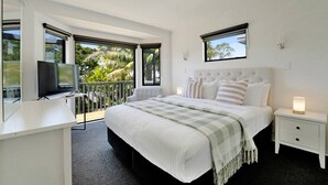 A modern bedroom with a large bed, a TV, and a balcony with a view of palm trees. - Seaside Premium Home with Spa Pool near Beach (Auckland East)