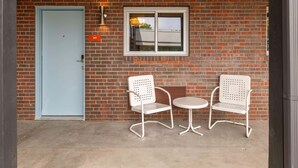 A brick building with a light blue door, two white metal chairs, and a small round table. - Desert Disco Dreams | La Vista Motel | 103 (Denver)
