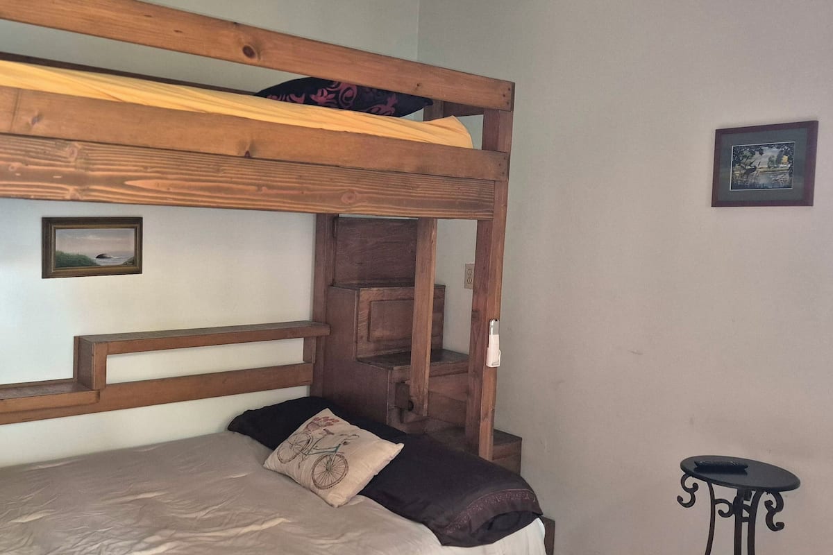 A bunk bed with a wooden headboard, a small bedside table, a framed picture on the wall, and a window with a view.