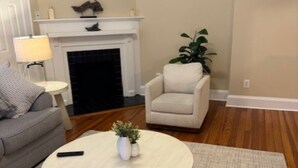 A living room with a fireplace, a sofa, a chair, a small table with a lamp, and a potted plant.