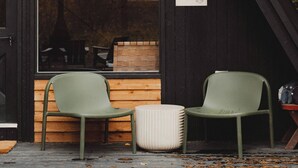 Eine Holzhütte mit geneigtem Dach, zwei grüne Stühle, ein weißer Hocker und ein kleiner Tisch.
