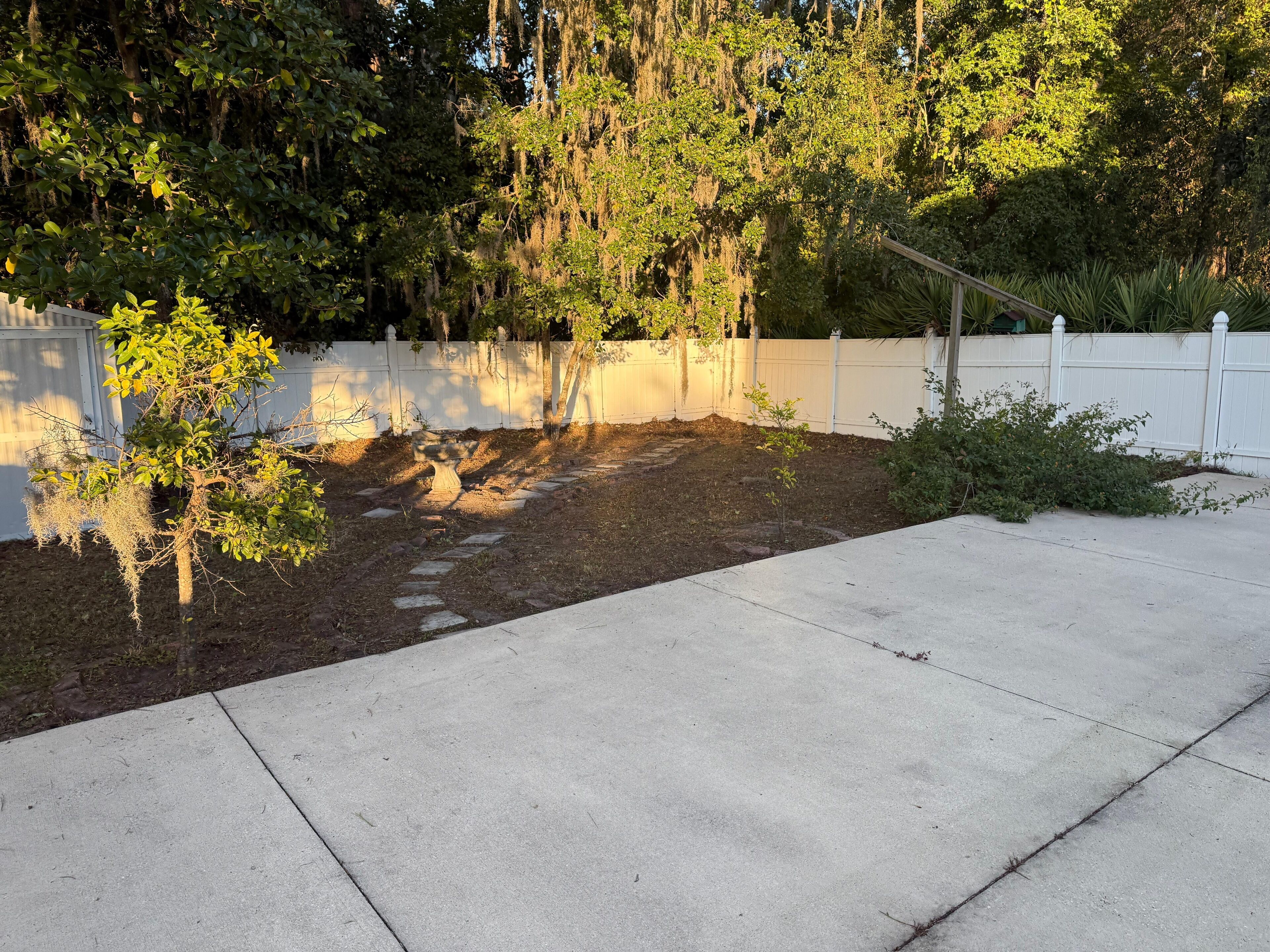 A backyard with a concrete patio, a small tree, and a white fence.