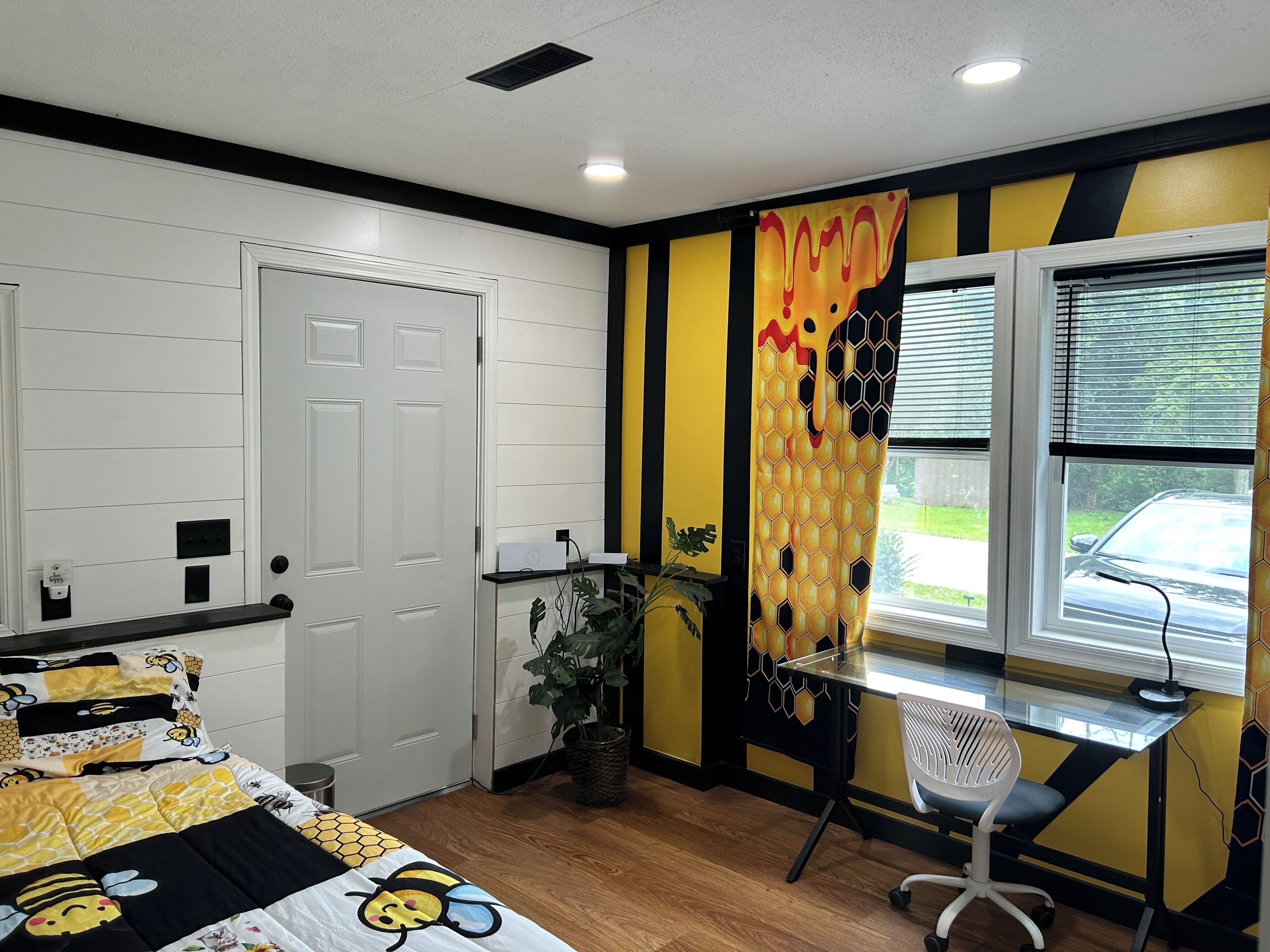 A bedroom with a bed, a desk, a chair, a window with blinds, and a door.