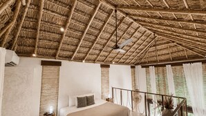 A bedroom with a thatched roof, a bed with a beige blanket, a nightstand, and a view of greenery through large windows. - Ikal Bungalows: HA is a poolside bungalow with AC in a jungle setting of Tulum. (Tulum)