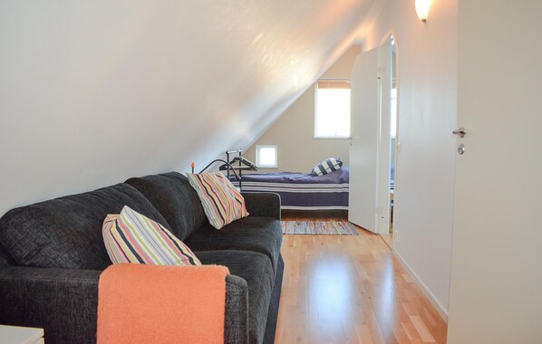 A cozy room with a dark grey sofa, a striped pillow, and a small orange throw blanket. A bed with a checkered pillow is visible in the background.